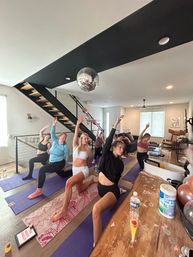 Group of people practicing yoga in a modern living-room home studio, lunging on colorful mats beneath a disco ball near a staircase.
