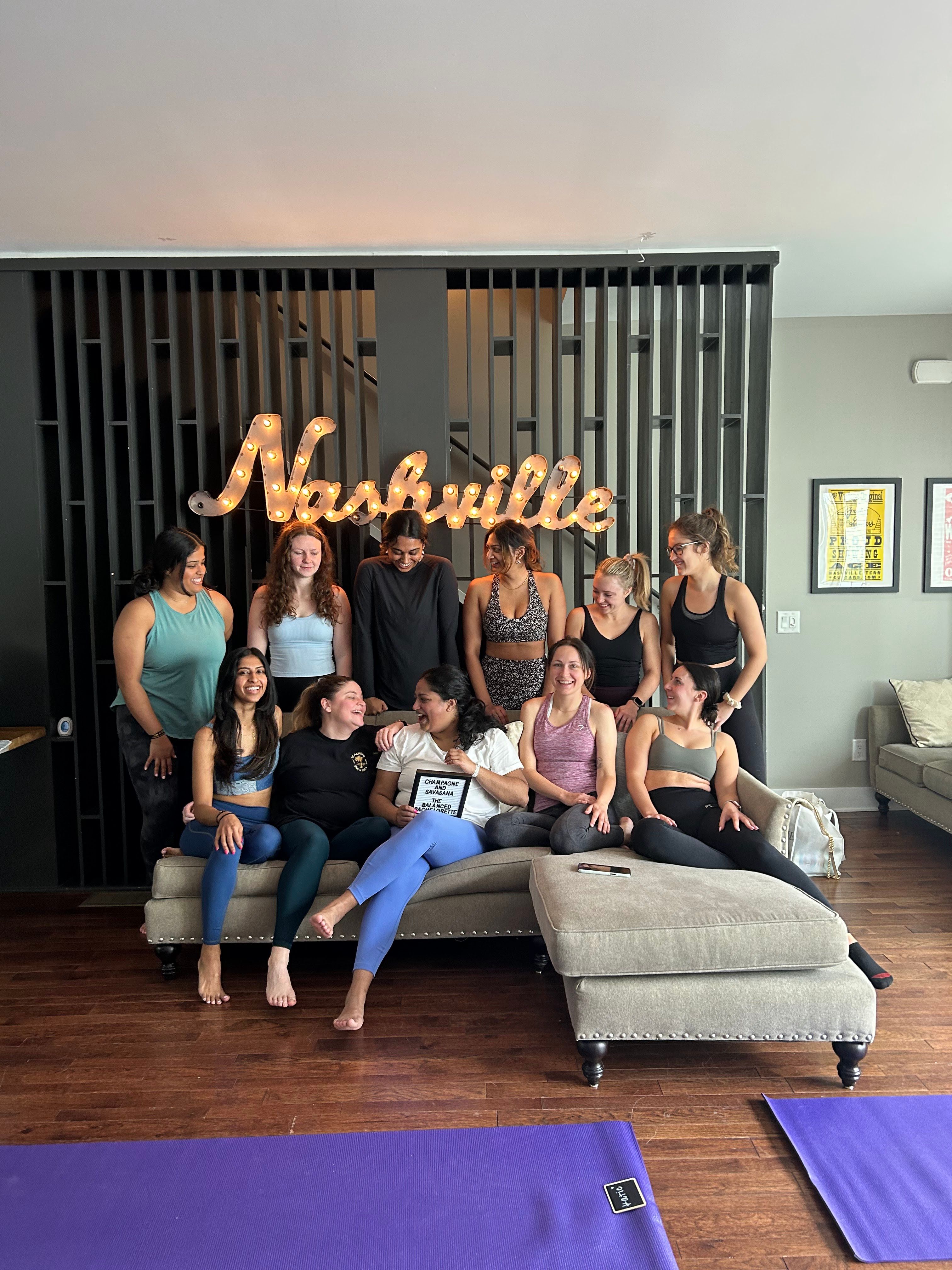 Smiling group of women in activewear gathered on a sofa under a lit "Nashville" marquee sign with purple yoga mats on a hardwood floor — friendly Nashville yoga class.