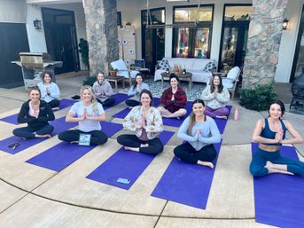 Smiling group of women seated in prayer pose on purple yoga mats during an outdoor backyard patio yoga class in front of a stone-column house and cozy lounge area.