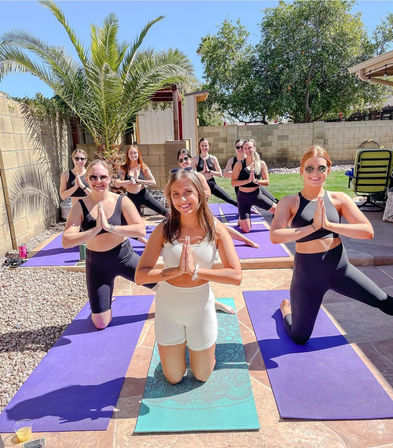 Outdoor backyard yoga class with a group of women kneeling on purple mats in prayer pose, smiling on a sunny patio under a palm tree