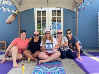 Five women smiling on yoga mats under a canopy on a blue patio for a bachelorette yoga party; center woman holds a balanced bachelorette sign, champagne balloon overhead and two strawberry-topped mimosas in the foreground.