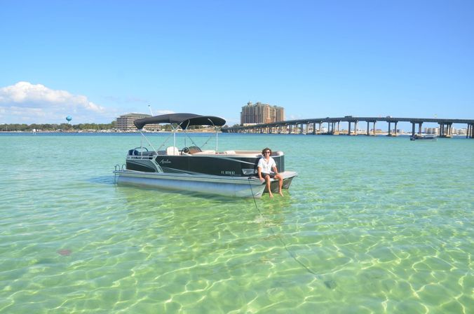 Person sitting on a pontoon boat anchored in shallow crystal-clear turquoise water with a long coastal bridge and beachfront buildings under a bright blue sky