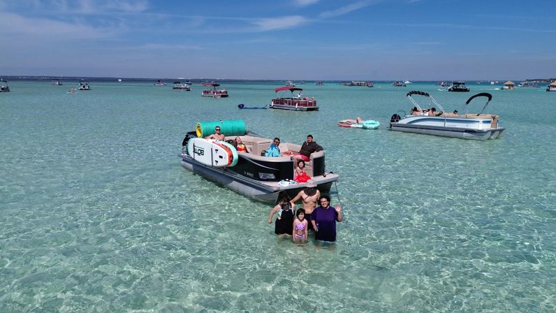 Group of people enjoying a pontoon boat anchored in shallow, crystal-clear turquoise water near a sandbar, with other boats and float tubes under a sunny blue sky.