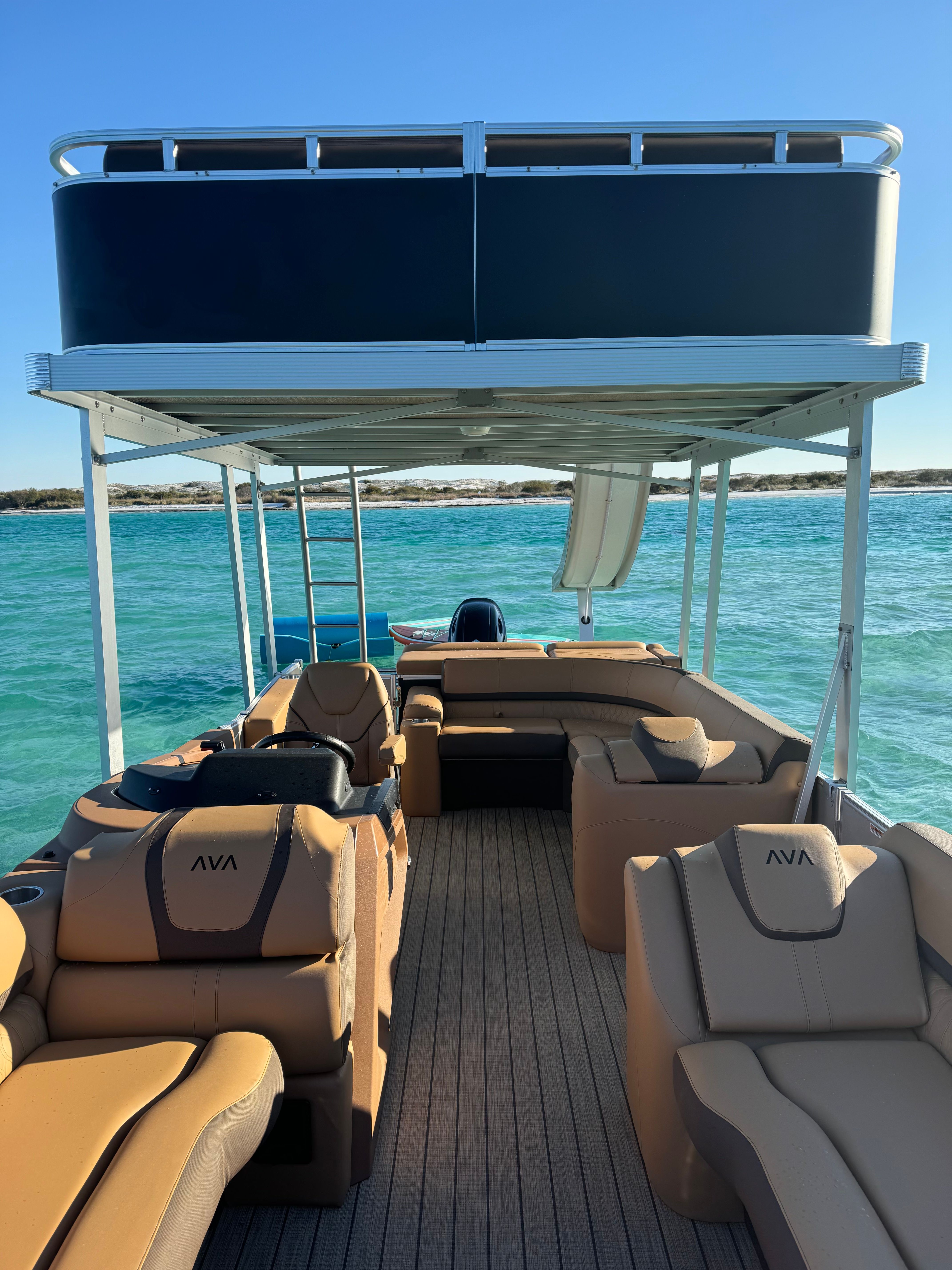 Interior view of a two-level pontoon boat with tan cushioned lounges, upper sun deck and slide, cruising on clear turquoise coastal waters with a sandy shoreline and bright blue sky