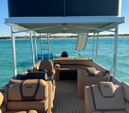 Interior view of a two-level pontoon boat with tan cushioned lounges, upper sun deck and slide, cruising on clear turquoise coastal waters with a sandy shoreline and bright blue sky