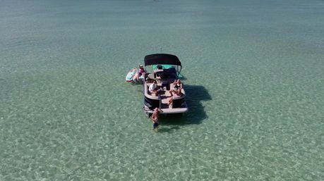 Aerial view of a sun-soaked pontoon boat anchored in crystal-clear shallow turquoise water, people lounging onboard with a paddleboard tied alongside and one person wading in the water.