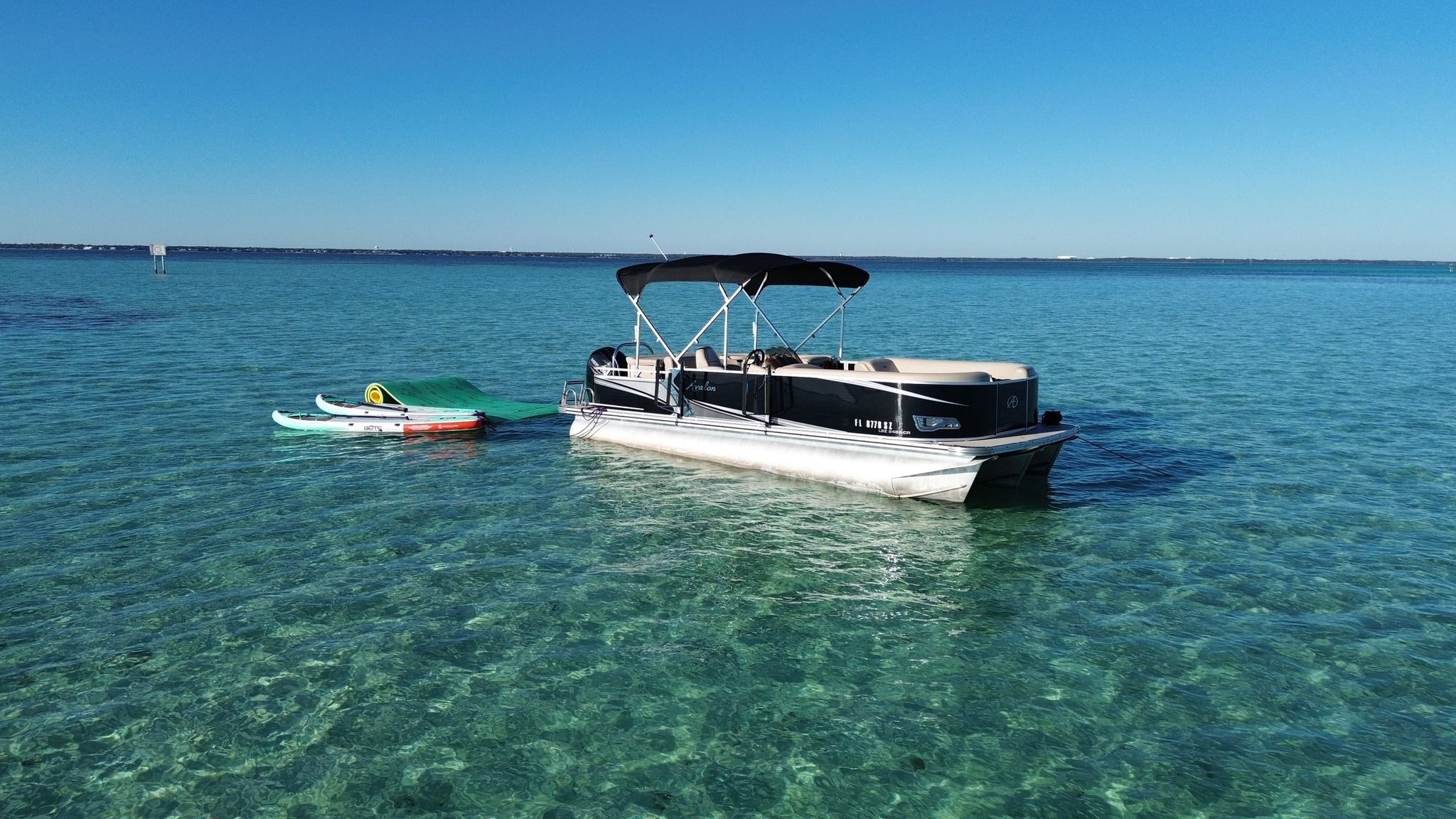 Pontoon boat anchored in crystal-clear turquoise coastal waters with two paddleboards and a green floating mat on a sunny, calm day.
