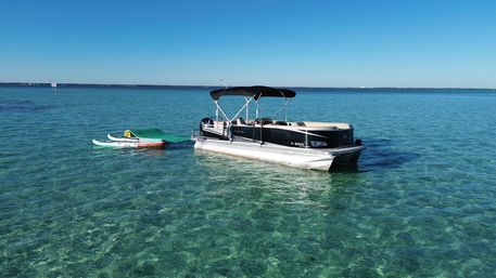 Pontoon boat anchored in crystal-clear turquoise coastal waters with two paddleboards and a green floating mat on a sunny, calm day.