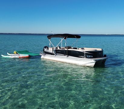 Pontoon boat anchored in crystal-clear turquoise coastal waters with two paddleboards and a green floating mat on a sunny, calm day.