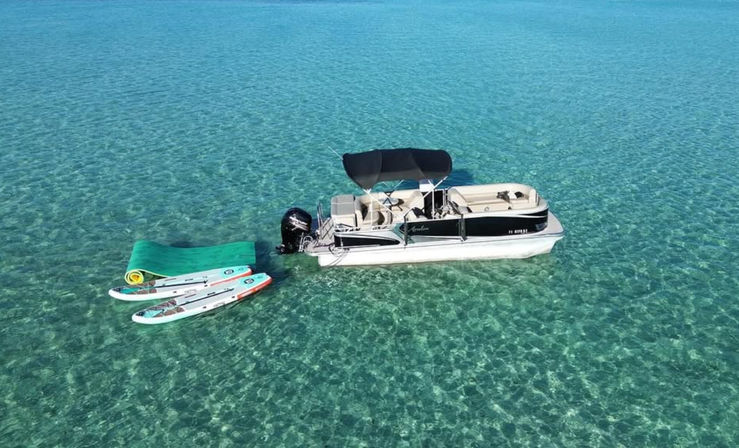 Pontoon boat with black canopy anchored in crystal-clear turquoise shallow water beside two stand-up paddleboards and a green floating mat on a sunny, calm day.