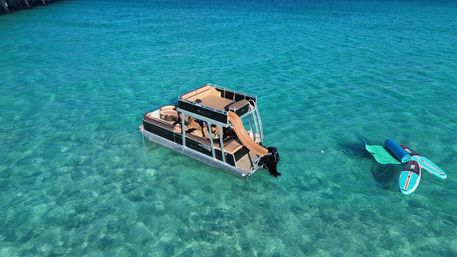 Aerial view of a modern pontoon boat with a tan slide anchored in crystal turquoise shallow water beside two colorful stand-up paddleboards and a floating mat.