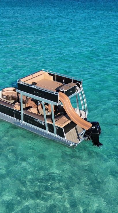 Aerial view of a modern pontoon boat with a tan slide anchored in crystal turquoise shallow water beside two colorful stand-up paddleboards and a floating mat.