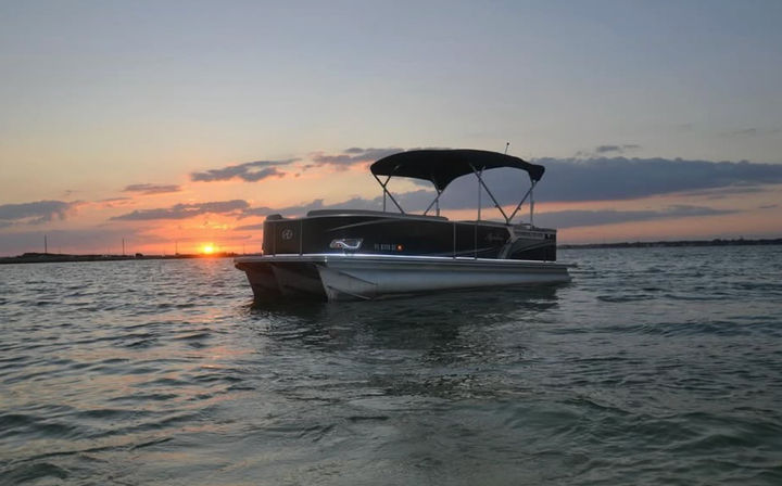 Pontoon boat floating on calm coastal water at sunset, vivid orange and pink sky reflecting on gentle waves with a distant shoreline — perfect sunset cruise scene