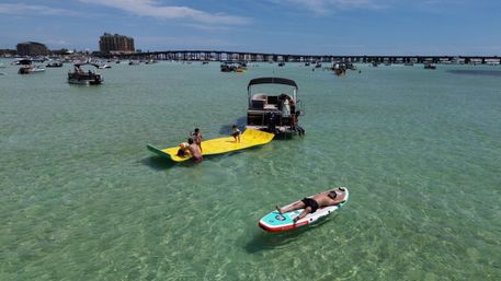Aerial view of a busy shallow turquoise bay with boats anchored near a long coastal bridge, families playing on a yellow floating mat and a man sunbathing on a paddleboard