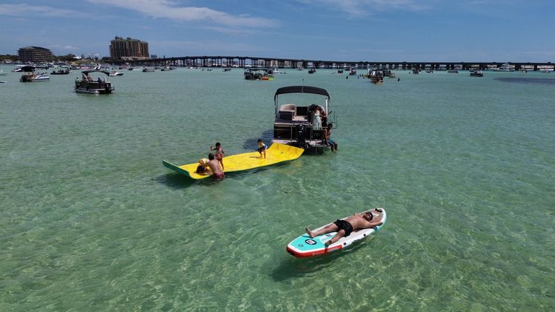 Aerial view of a busy shallow turquoise bay with boats anchored near a long coastal bridge, families playing on a yellow floating mat and a man sunbathing on a paddleboard
