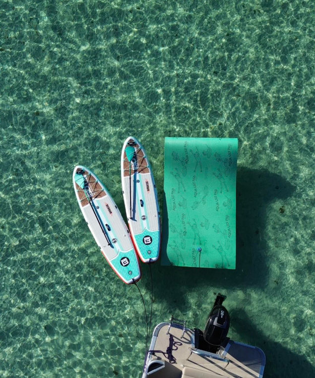 Aerial view of two turquoise-and-white stand-up paddleboards and a green inflatable floating mat tied to a boat swim platform, floating on sunlit crystal-clear shallow turquoise water.