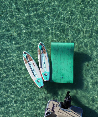 Aerial view of two turquoise-and-white stand-up paddleboards and a green inflatable floating mat tied to a boat swim platform, floating on sunlit crystal-clear shallow turquoise water.
