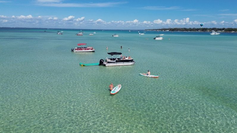 Aerial view of a shallow turquoise bay on a sunny day with anchored pontoon boats, people paddleboarding and kayaking in clear water near a tree-lined shoreline.