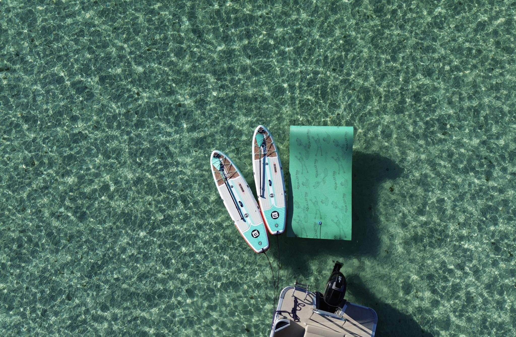 Aerial view of two stand-up paddleboards tied to a bright inflatable floating dock beside a small motorboat on crystal-clear turquoise water.