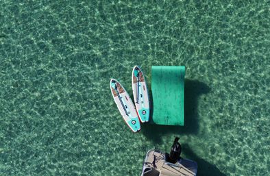 Aerial view of two stand-up paddleboards tied to a bright inflatable floating dock beside a small motorboat on crystal-clear turquoise water.