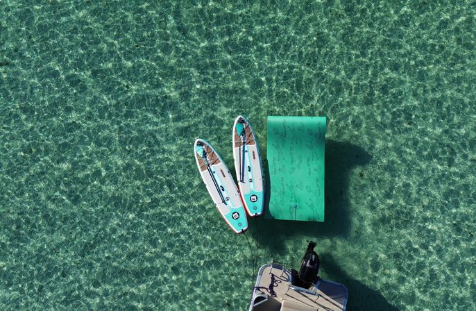 Aerial view of two stand-up paddleboards tied to a bright inflatable floating dock beside a small motorboat on crystal-clear turquoise water.
