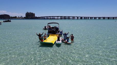Group waving from a pontoon boat with a yellow floating slide and paddleboards in clear turquoise shallow bay, bridge and resort skyline under a bright blue sky.