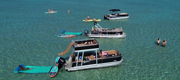 Pontoon boats anchored in turquoise, crystal-clear water with a slide, paddleboards and people relaxing and swimming on a sunny summer day