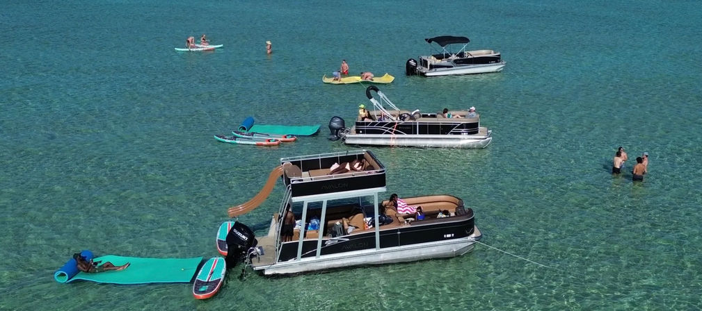 Pontoon boats anchored in turquoise, crystal-clear water with a slide, paddleboards and people relaxing and swimming on a sunny summer day