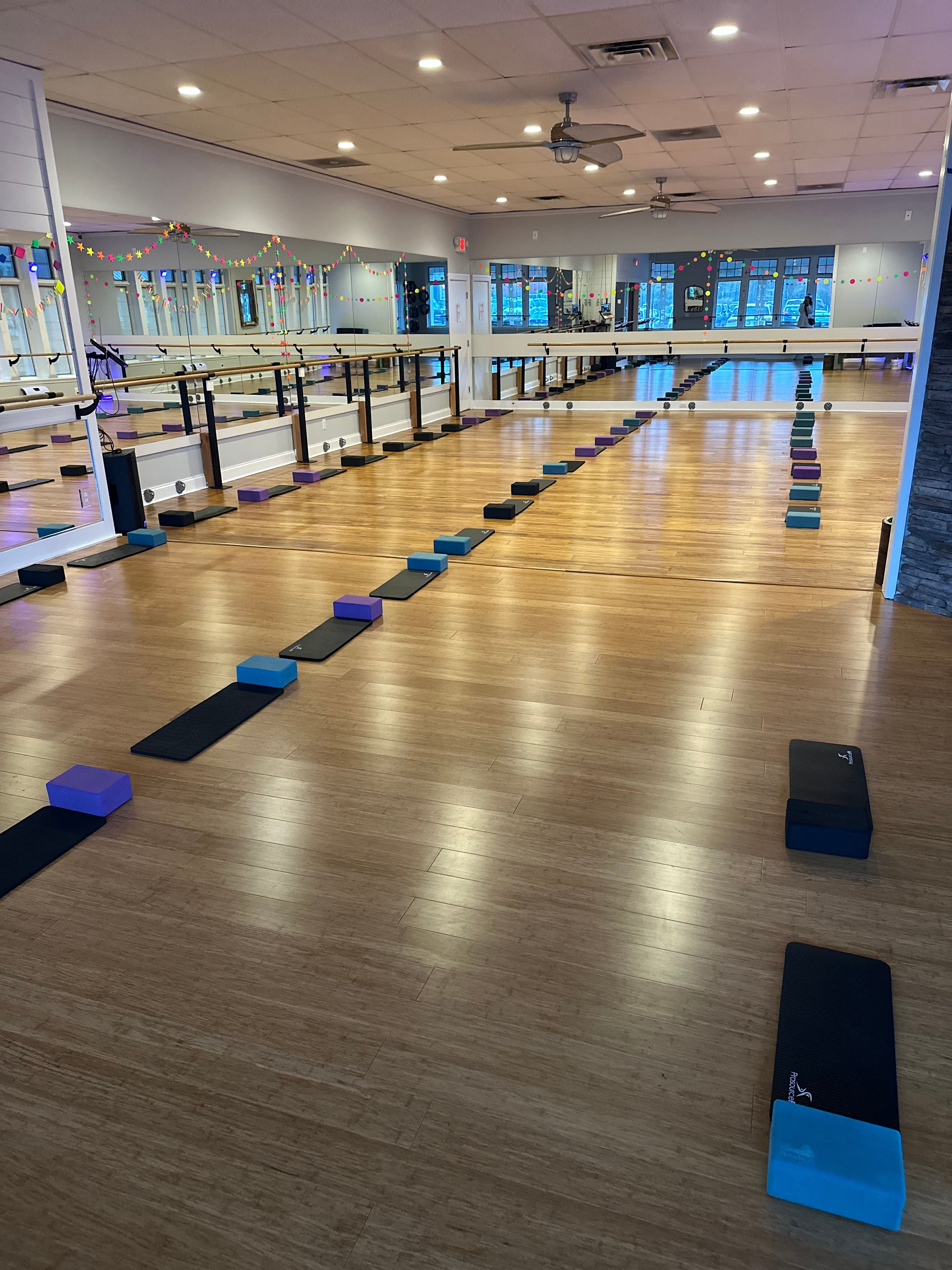 Spacious barre and yoga fitness studio with mirrored wall and wood floor, rows of black mats and colorful foam blocks neatly lined for class, wall barres, ceiling fans and decorative string lights visible.