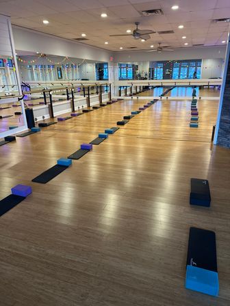 Spacious barre and yoga fitness studio with mirrored wall and wood floor, rows of black mats and colorful foam blocks neatly lined for class, wall barres, ceiling fans and decorative string lights visible.