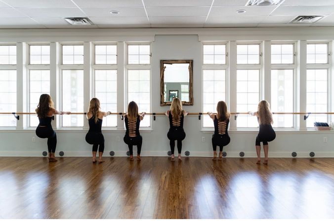 Six participants in black outfits doing barre squats at a wooden barre in a light-filled barre class studio with tall windows and hardwood floors.