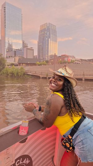 Smiling woman in a straw cowboy hat and yellow tank top leaning on a boat railing on a river at sunset, with reflective downtown skyscrapers and a riverfront promenade in the background