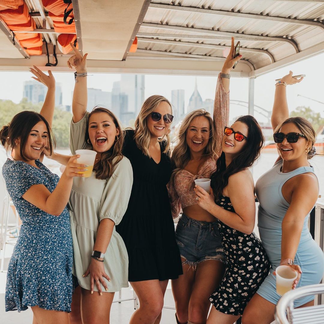 Six women in summer dresses and sunglasses cheering with drinks on a daytime riverboat cruise, city skyline and bridge visible in the background.