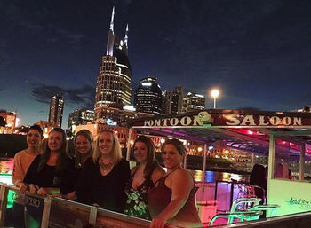 Group of friends on a lit pontoon boat at night with the Nashville skyline and illuminated skyscrapers reflecting on the river.