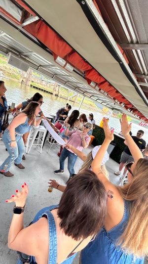 Daytime riverboat party on a covered deck with friends dancing, cheering and holding drinks near a docked river, casual summer outfits and sunglasses.
