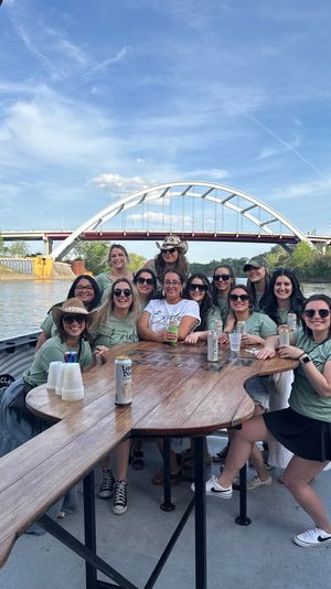 Group of women on a river cruise boat deck celebrating a bachelorette party around a wooden table, with an arched bridge and city skyline in the background under a blue sky.