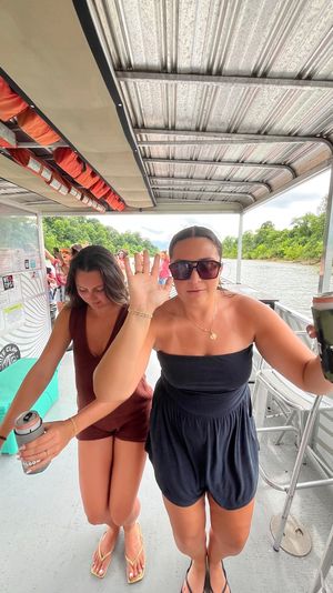 Two people on a pontoon boat on a tree-lined river in summer, one waving and holding a canned drink in a camouflage cooler, overhead life jackets, casual summer outfits and flip-flops, relaxed boat outing