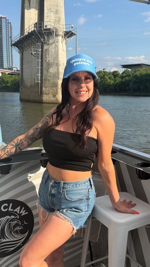 Smiling woman on a boat ride on an urban river, leaning by a concrete bridge pillar, wearing a blue cap that reads "spitters are quitters", black tube top, ripped denim shorts and a sleeve tattoo under a sunny sky with city skyline and trees in the background.
