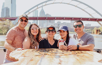 Cheerful group of five adults posing at a wooden table on a sunny riverboat, city skyline and arched bridge behind them; one person holding a canned drink.