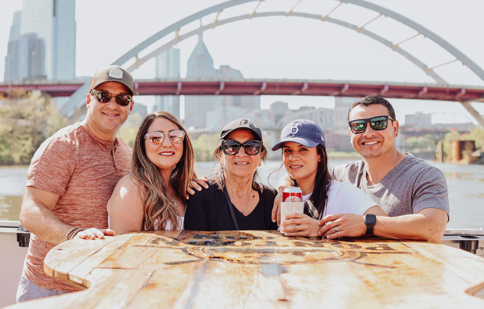 Cheerful group of five adults posing at a wooden table on a sunny riverboat, city skyline and arched bridge behind them; one person holding a canned drink.