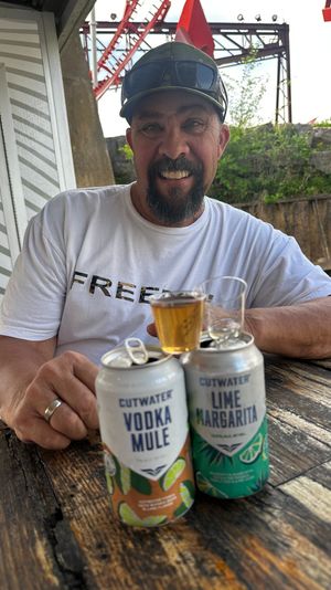 Smiling man in a cap at an outdoor waterfront wooden table with two Cutwater canned cocktails (Vodka Mule, Lime Margarita) and a small pour, red roller coaster track and greenery in background.