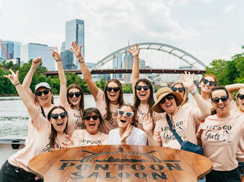 Cheerful group of women in matching pink "Let's Go Girls" shirts and sunglasses waving and laughing on a river pontoon boat with an arched bridge and downtown skyline in the background — fun downtown river cruise scene.
