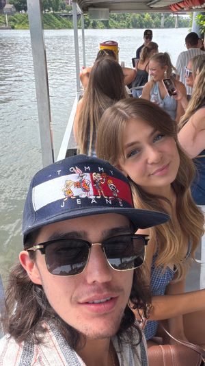 Smiling friends taking a selfie on a sunny riverboat cruise, man in sunglasses and patterned cap in the foreground with waterfront and tree-lined shore in the background.