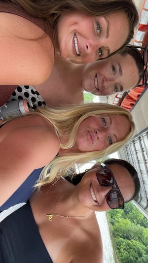Four smiling friends in swimsuits taking a close-up selfie on a boat with a tree-lined lake in the background, sunny summer vibe