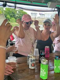 Women cheering on a riverside pontoon boat party, wearing pink caps and heart-shaped sunglasses, raising hands over a wooden table with canned drinks in colorful koozies, green trees and shoreline tanks in the background.