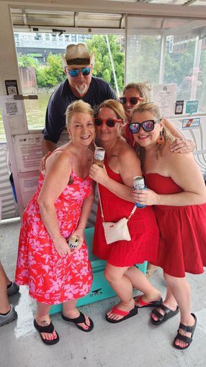 Cheerful group on a sunny riverboat cruise: four women in red dresses and sunglasses holding canned drinks with a man in a cowboy hat and mirrored shades behind them, river and waterfront buildings visible through the windows.