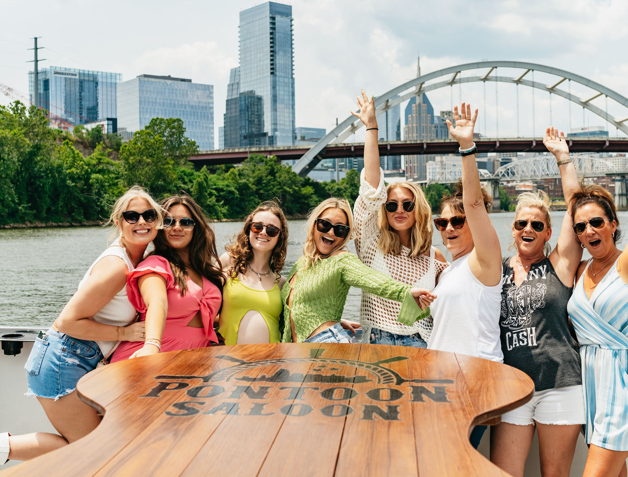 Eight women in sunglasses celebrating on a pontoon boat on the Cumberland River with the Nashville skyline and an arched bridge in the background, sunny summer vibes