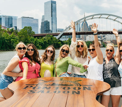 Eight women in sunglasses celebrating on a pontoon boat on the Cumberland River with the Nashville skyline and an arched bridge in the background, sunny summer vibes