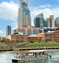 Small passenger pontoon boat cruising the Cumberland River with people aboard, passing Nashville downtown skyline of modern skyscrapers and red‑brick riverfront buildings on a sunny day.