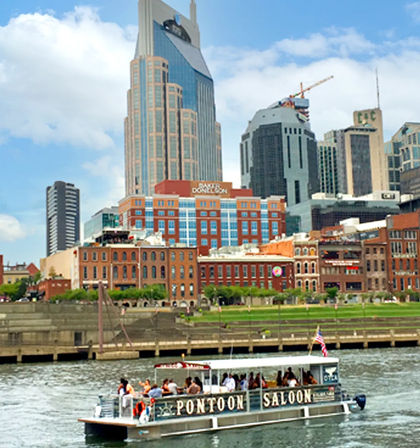Small passenger pontoon boat cruising the Cumberland River with people aboard, passing Nashville downtown skyline of modern skyscrapers and red‑brick riverfront buildings on a sunny day.